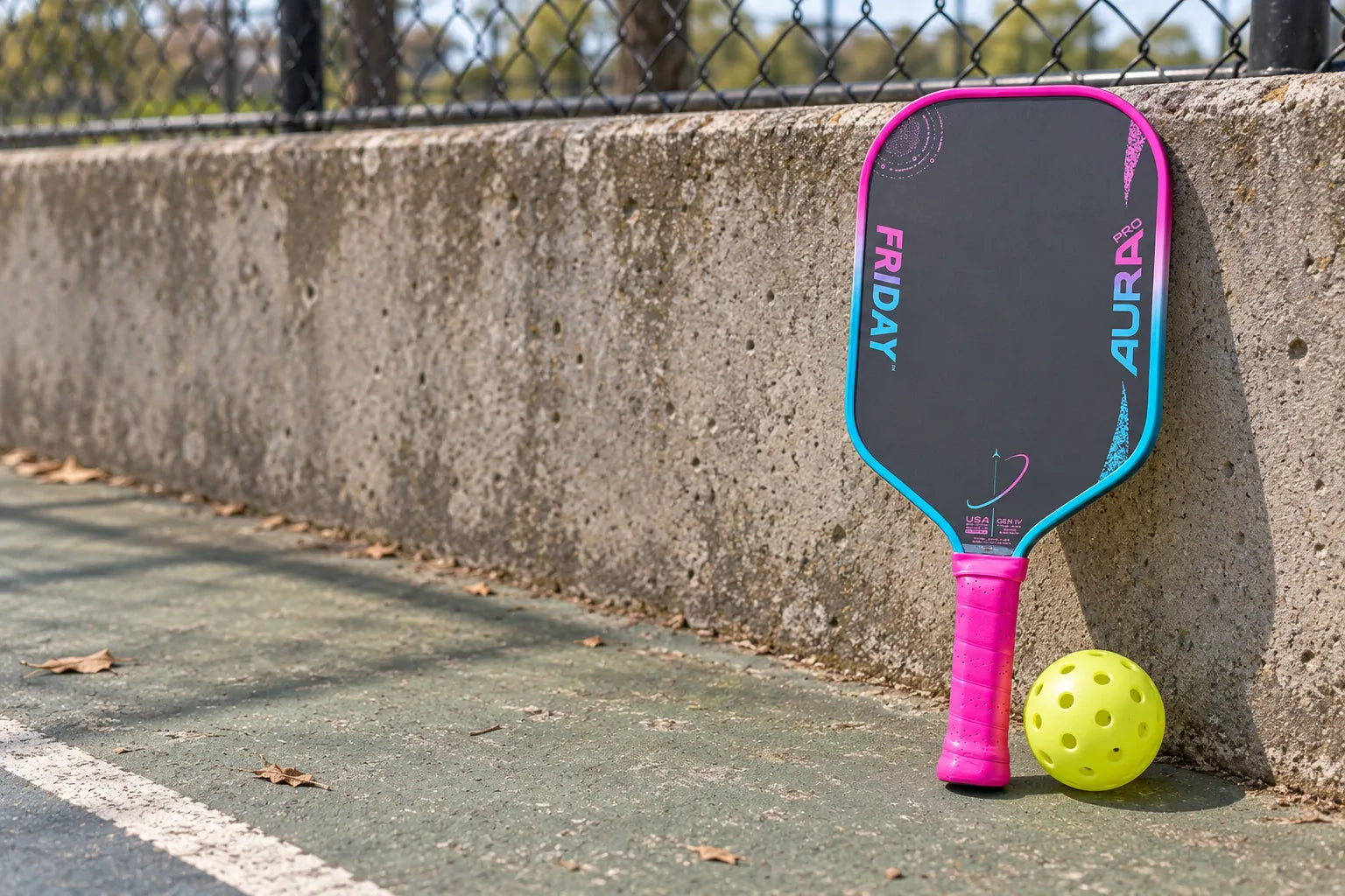 Friday Aura Pro paddle and a pickleball against a courtside wall